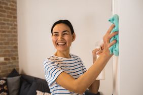 Woman wearing a white t-shirt with blue stripes smiling while she cleans a wall with a spray bottle and blue cloth