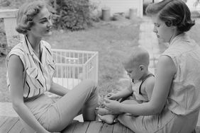 Two women sit on the front porch and visit over a cup of coffee. One of the women, a mother holds her child, a toddler boy, in her lap