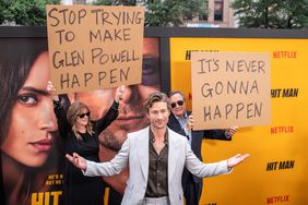 Glen Powell stands in front of his parents holding up signs making fun of him.