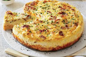 A bacon and cheddar grits quiche sits on a table with one slice cut out.