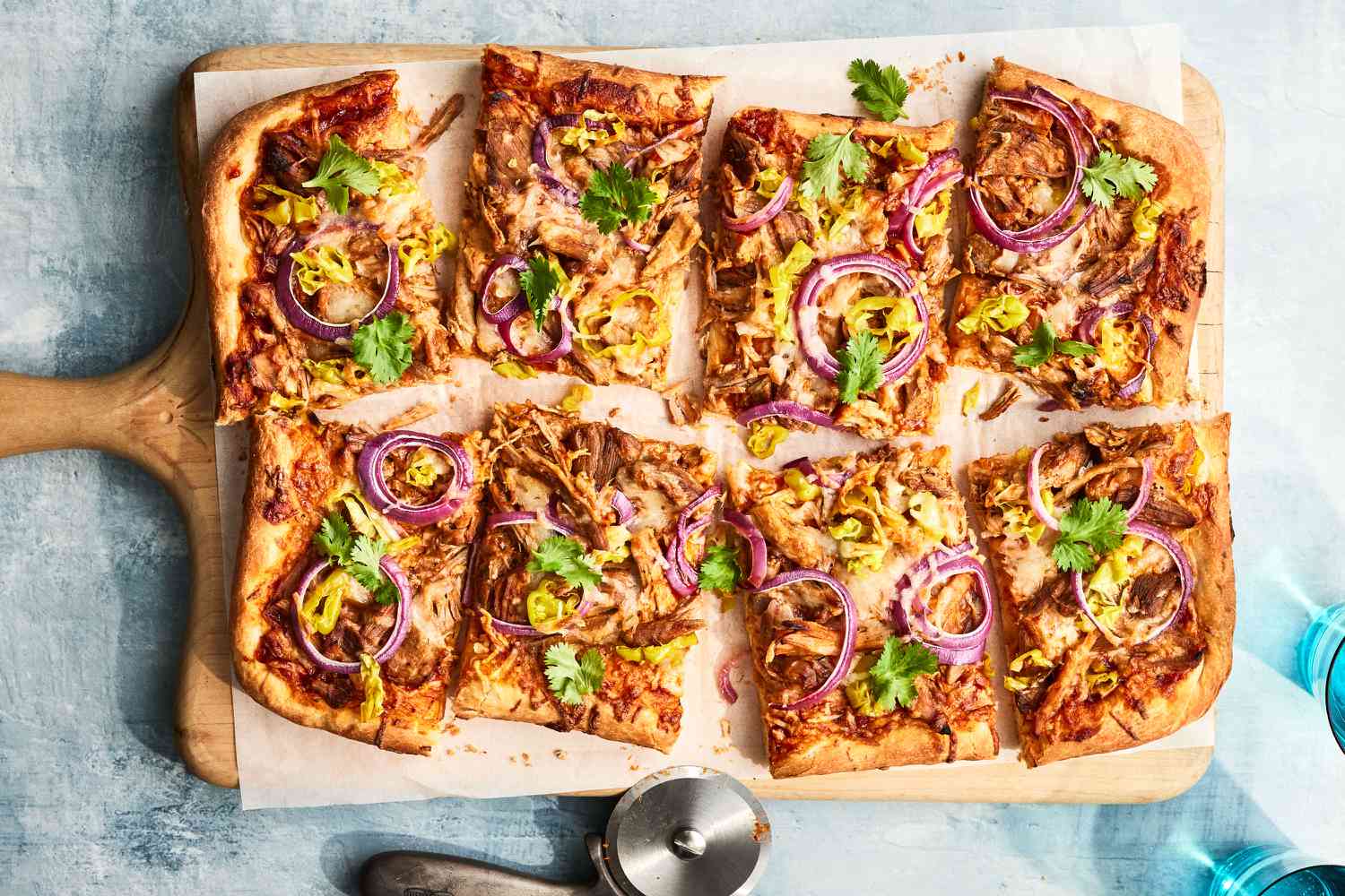 Cut barbecue pork pizza on a cutting board on a table with a pizza cutter and two glasses of water beside.